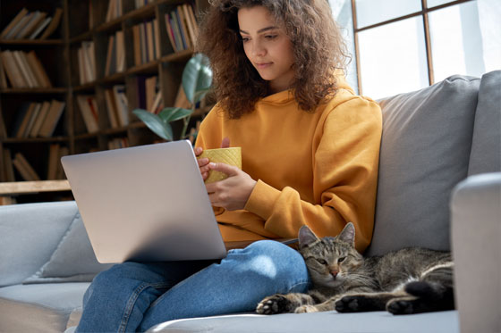 Young female sitting on a sofa using a laptop with a cat by her side.